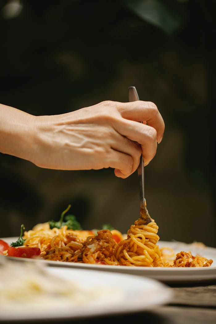 Crop faceless female with fork eating freshly cooked scrumptious Bolognese pasta served on white ceramic plate